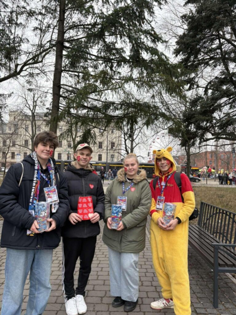 Członkowie zespołu projektowego „Podaruj radość - jest bezcenna” kwestują na rzecz Wielkiej Orkiestry Świątecznej Pomocy. Stoją na świeżym powietrzu, trzymając puszki WOŚP i identyfikatory, a jedna osoba jest przebrana w strój Kubusia Puchatka.